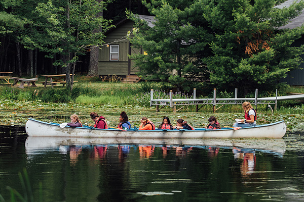 A group of people wearing life vests sit in a large canoe on a lake