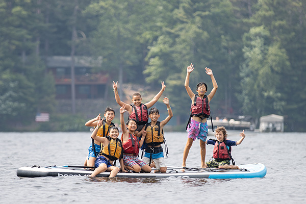 A group of children wearing life vests waving on a large paddleboard on a lake