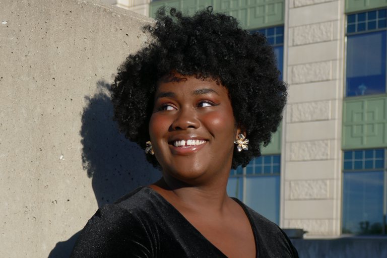 A black female student who is wearing a black top and standing outside in front of part of a concrete wall and a building smiles and looks off to the side.