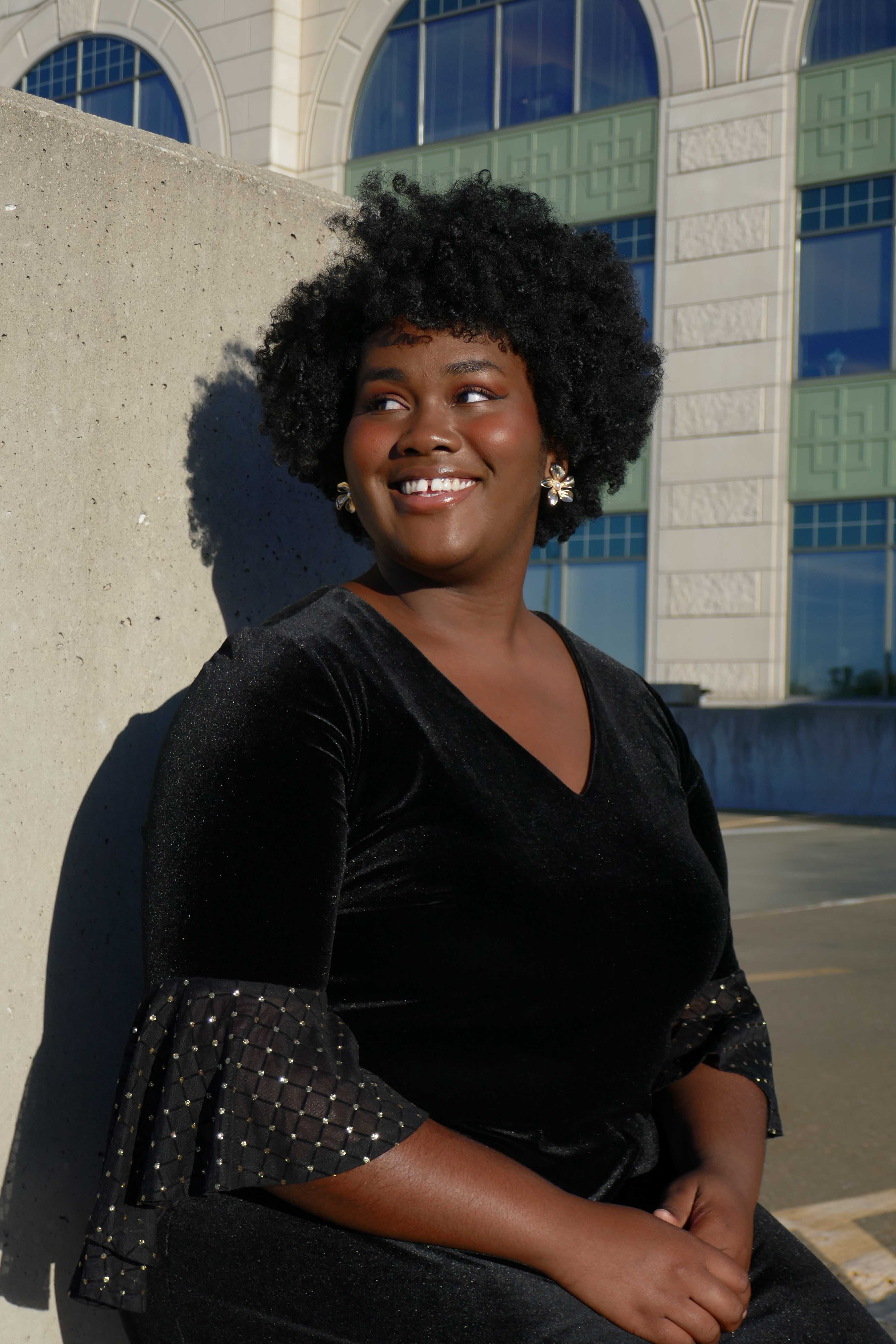 A black female student who is wearing a black top and standing outside in front of part of a concrete wall and a building smiles and looks off to the side.