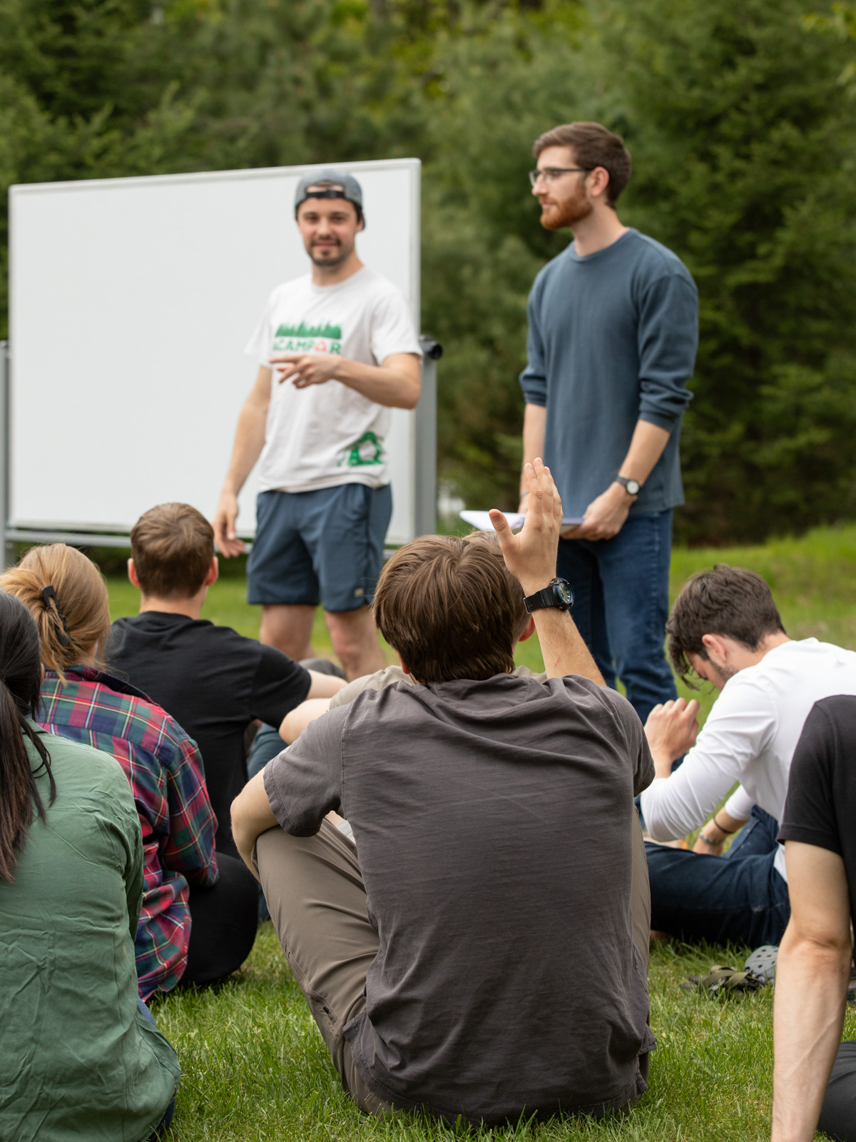 A group of young adults sit on grass while listening to two men speak