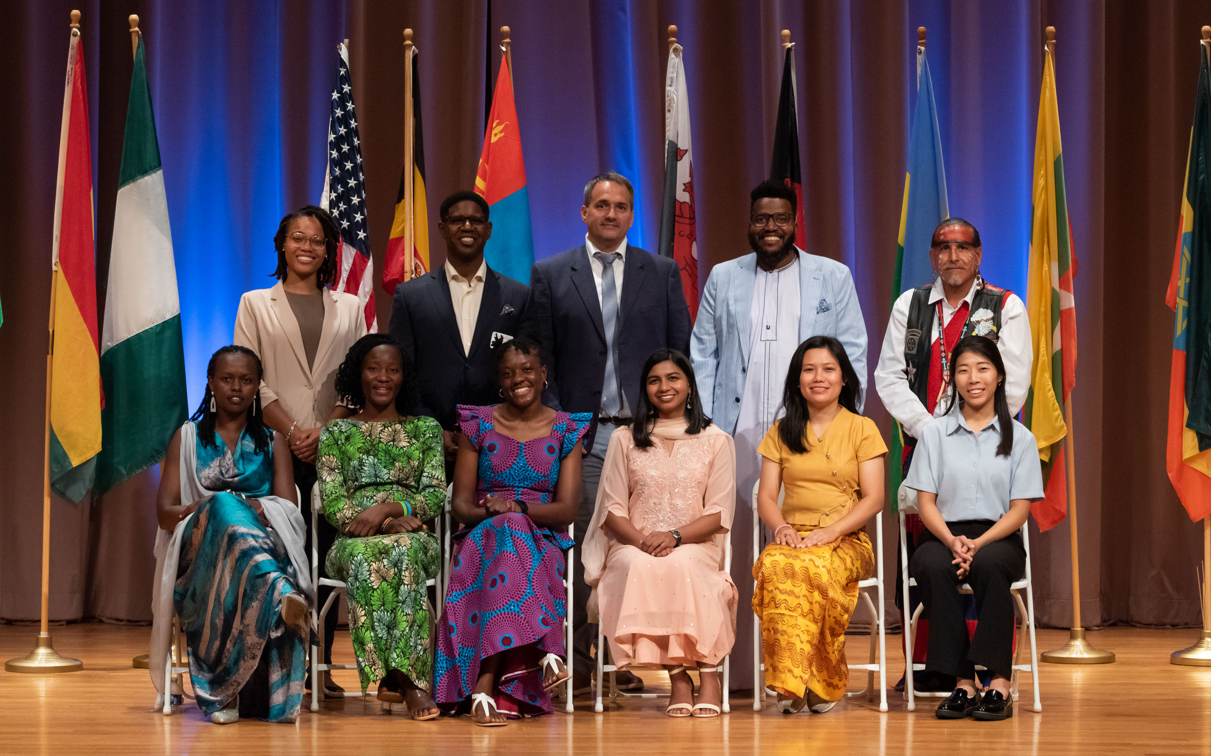 A group of eleven adults from various countries wear traditional clothing from their country and smile for a photo