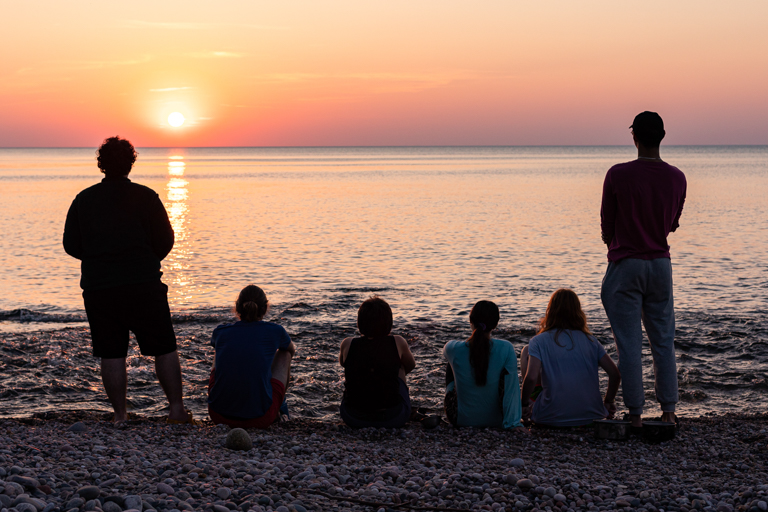 Six young adults on a beach look at a sunset