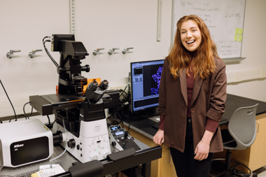 A smiling woman with ginger hair stands to the right of a microscope in a lab.