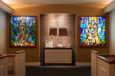 The front of chapel which contains a large tan cross hanging on the brick wall in between two stain glass windows. In front of the cross lies a table containing a few items, including an open Bible.