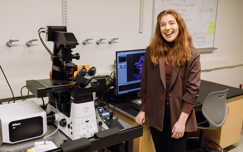 A smiling woman with ginger hair stands to the right of a microscope in a lab.