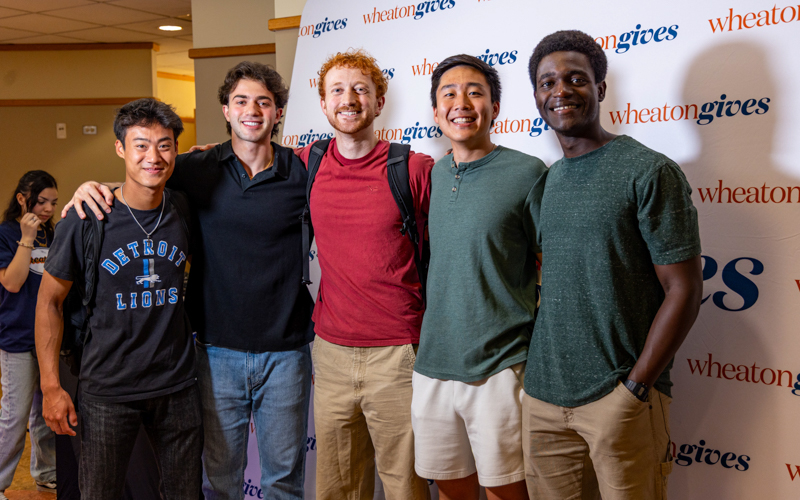 Five young male students smile and stand in front of a backdrop that says Wheaton Gives