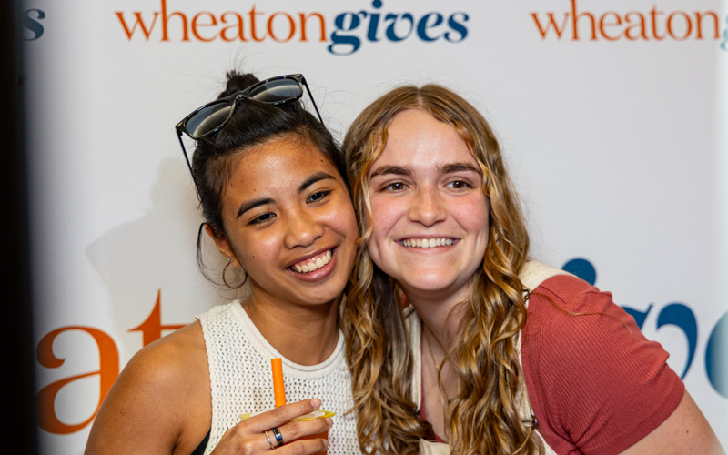 An east asian young woman and a white young woman smile in front of a backdrop that says Wheaton Gives